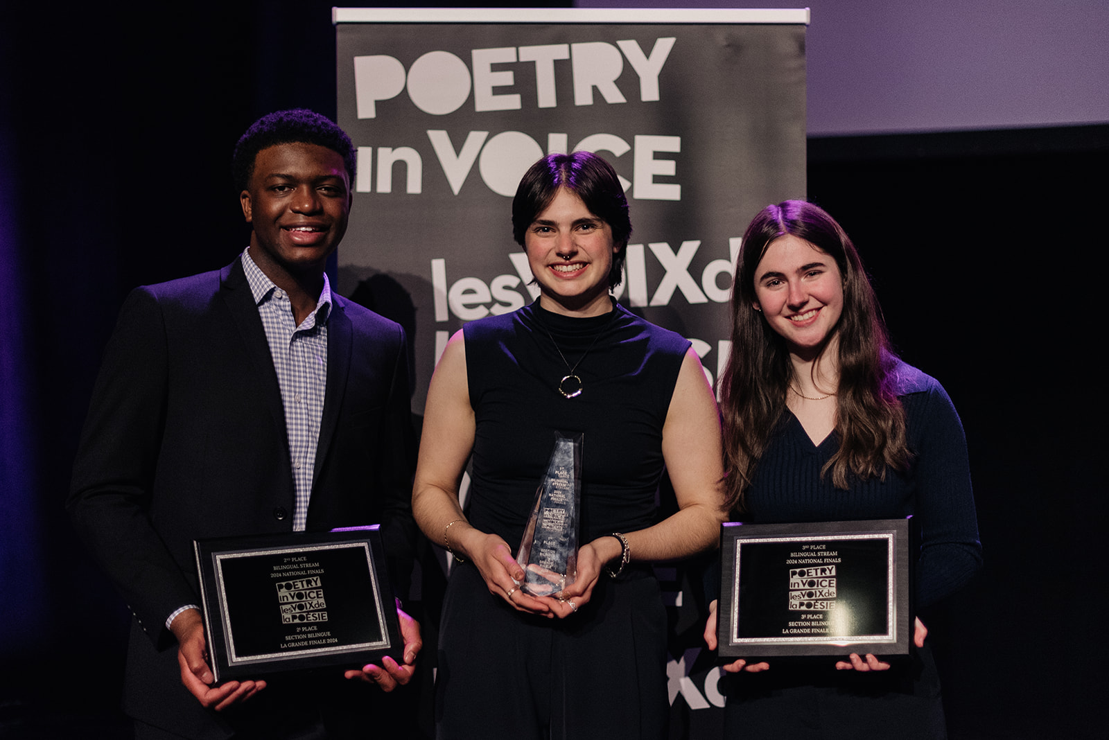 Three student recitation winners in the Bilingual stream smiling and holding their commemorative trophies and plaques in front of the Poetry in Voice banner.