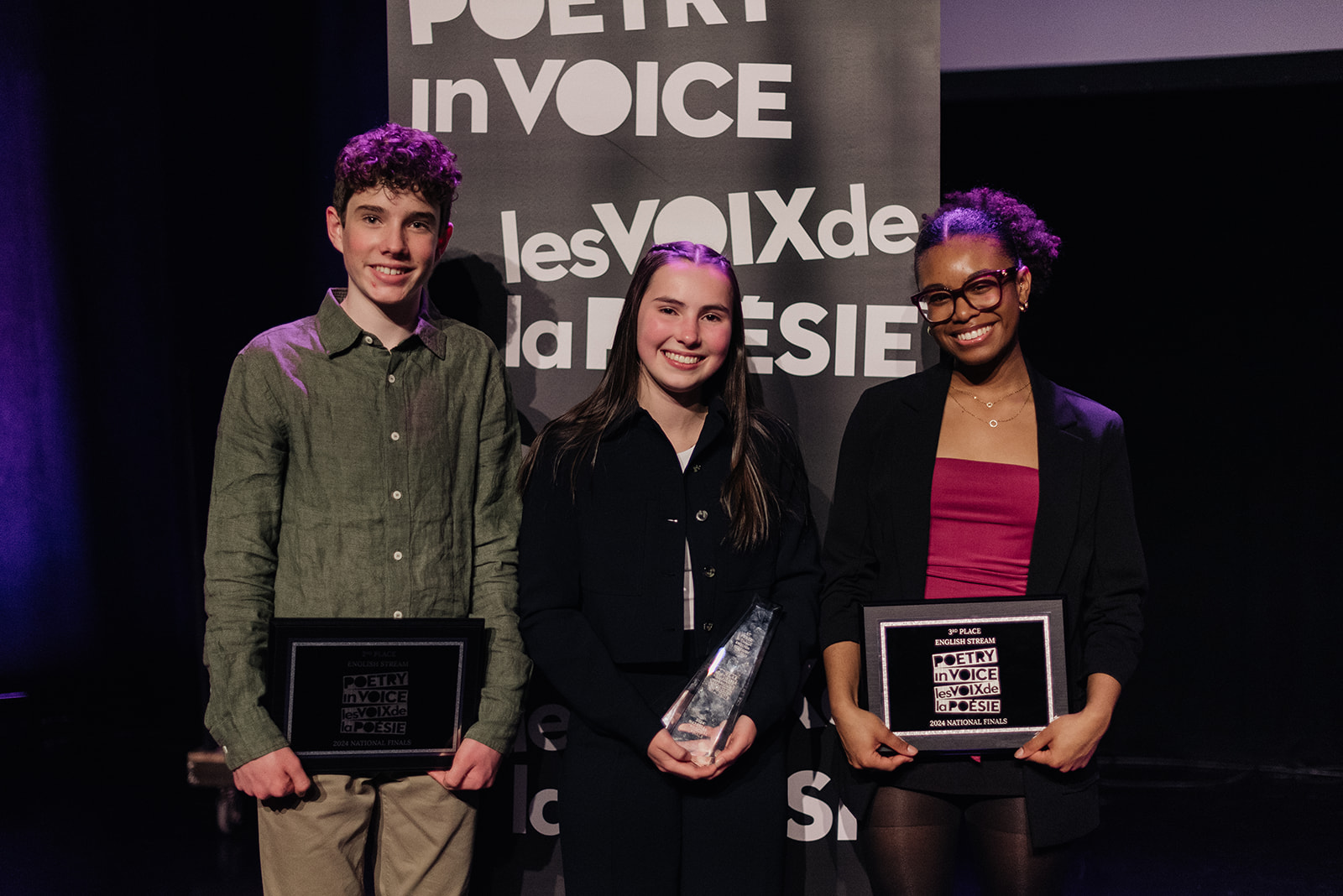 Three student recitation winners in the English stream smiling and holding their commemorative trophies and plaques in front of the Poetry in Voice banner.