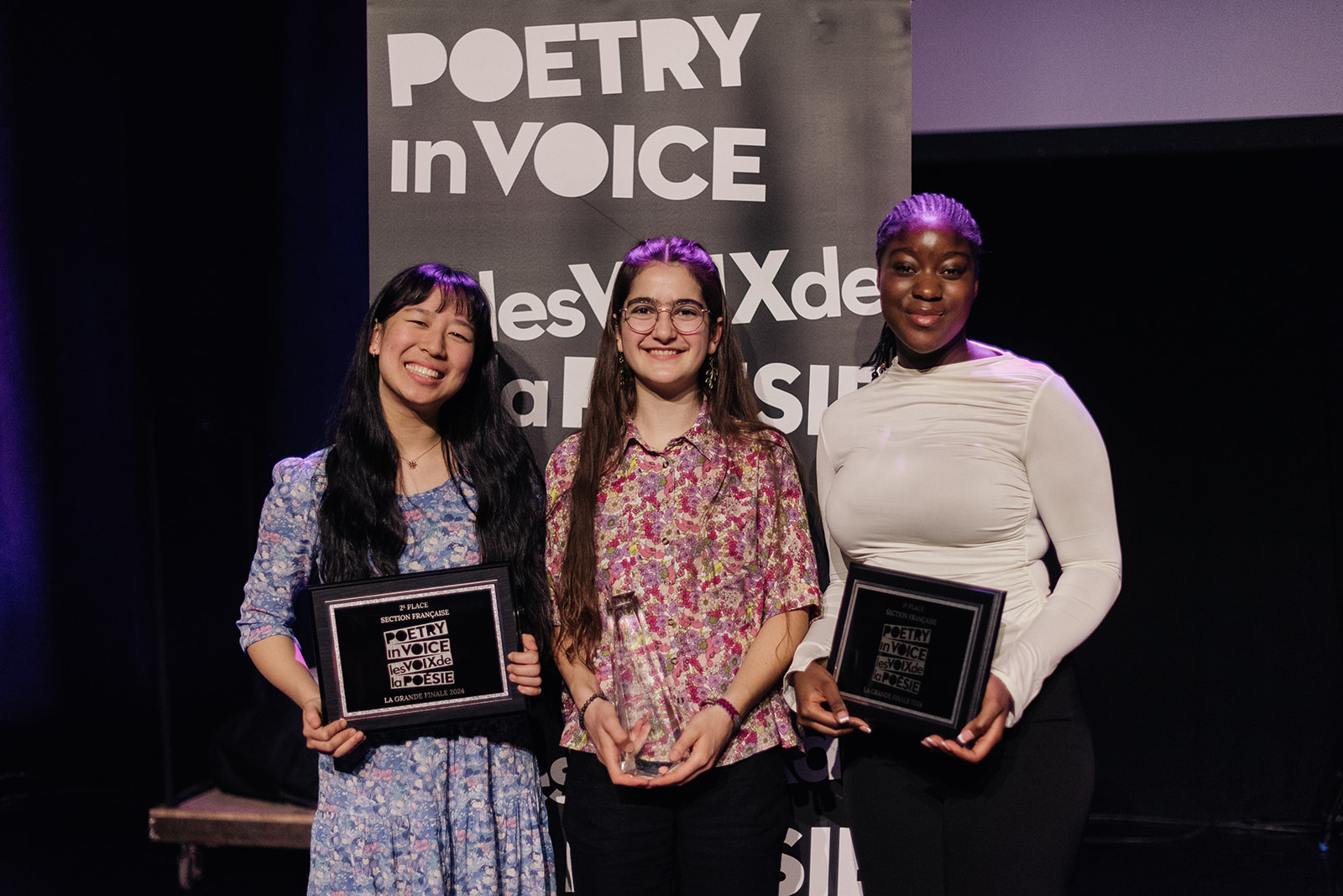 Three student recitation winners in the French stream smiling and holding their commemorative trophies and plaques in front of the Poetry in Voice banner.