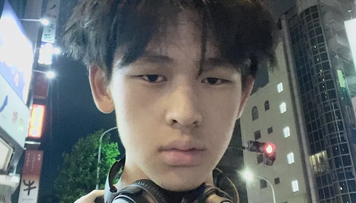 A young man in a city with headphones around his neck looks directly at the camera