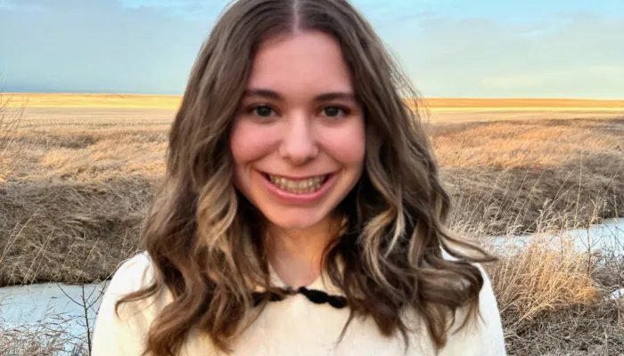 Photo of a young white girl in a white short sleeve knit shirt. She has brown hair and is smiling as she stands in a field against a wide blue sky.